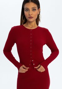 Czerwony kardigan w prążki z długimi rękawami, okrągłym dekoltem i zapinanym przodem na guziki. Posiada błyszczące srebrne guziki wzdłuż zapięcia.