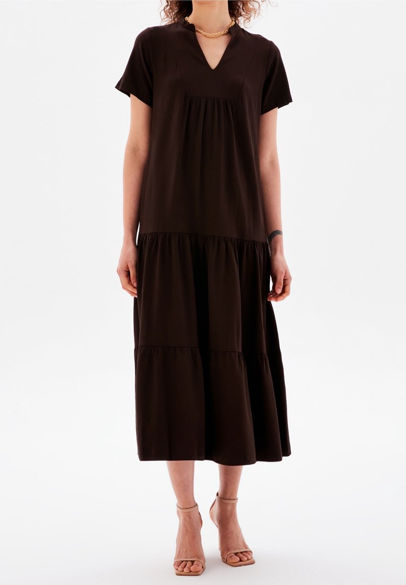 Femme portant une robe midi ample à manches courtes, à volants, marron foncé, avec un décolleté en V et des sandales à talons beiges.