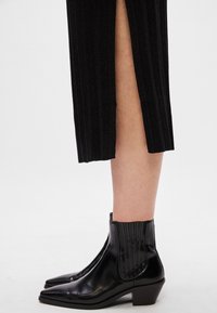 Cizme negre din piele până la gleznă, cu vârf ascuțit, panouri laterale elastice cu nervuri și toc mic tip block, asortate cu o fustă plisată neagră.