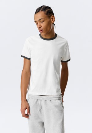 Jeune homme aux cheveux tressés portant un t-shirt blanc avec un col noir et des bordures de manches noires, ainsi qu'un pantalon de survêtement gris clair, posant les mains dans les poches.