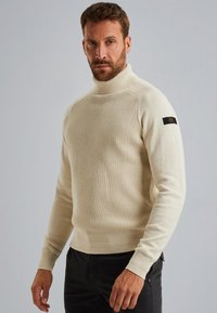 Hellbeige gestrickter Pullover mit hohem Kragen und strukturiertem Muster; besitzt gerippte Bündchen und ein Logopatch am linken Ärmel.