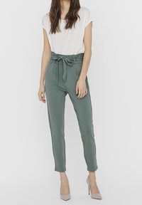 Pantalons joggers verts taille haute avec ceinture à nouer, poches latérales, jambes fuselées et texture douce. Portés avec un haut à manches courtes blanc et des talons.