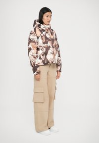 Giacca a vento marrone, bianca e rosa con un motivo geometrico animale. Abbinata a pantaloni cargo beige e sneakers bianche.