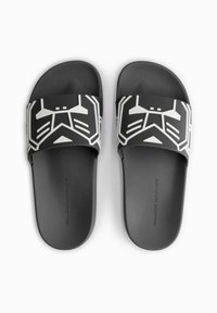 Black slide sandals with a wide strap featuring a white geometric pattern resembling a mask. Textured footbed for grip and comfort.