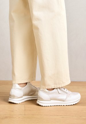 Beige bukser med vide ben kombineret med hvide teksturerede snørebåndssneakers, der står på trægulv.