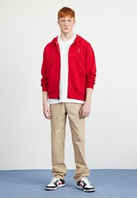 Red zip-up hoodie in cotton, white t-shirt, beige trousers, and black and white sneakers with red accents. Casual, sporty ensemble.
