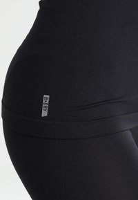 Detailní záběr na osobu v černých sportovních legínách a černém topu s malou etiketou "B-DRY" na boku poblíž pasu.