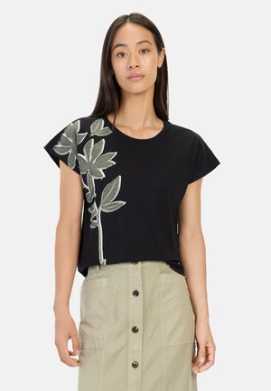 Zwarte t-shirt met groene bladeren borduurwerk aan één kant, korte mouwen en een relaxte pasvorm. Gepaarde met een beige knopenrok.
