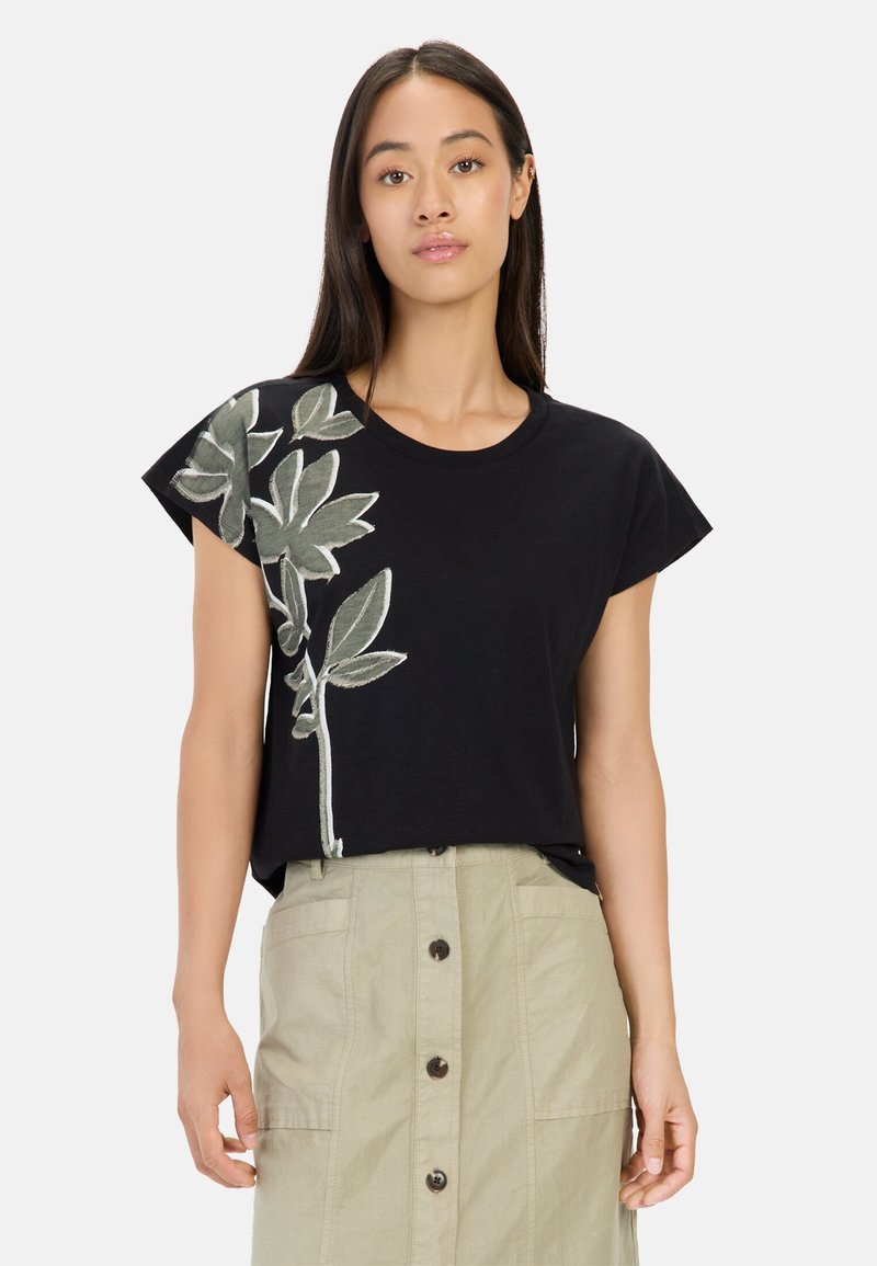 Zwarte t-shirt met groene bladeren borduurwerk aan één kant, korte mouwen en een relaxte pasvorm. Gepaarde met een beige knopenrok.