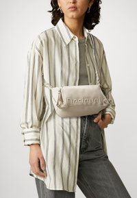 Beige Crossbody-Tasche aus Kunstleder mit Prägung, silberner Hardware und subtiler Textur, kombiniert mit einem gestreiften Hemd und grauen Jeans.