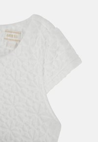 Camiseta blanca texturizada con un patrón floral, mangas cortas y cuello redondo. Presenta una pequeña etiqueta "GUESS" cerca del cuello.