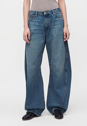 Personne portant un jean bleu taille large à jambes larges avec des coutures visibles, associé à des chaussures noires et un débardeur gris, debout devant un fond blanc.