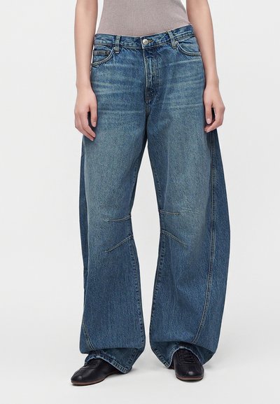 Persona que lleva vaqueros azules de corte ancho y caída suelta con costuras visibles, combinados con zapatos negros y una camiseta sin mangas gris, de pie frente a un fondo blanco.
