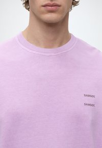Νεαρό άτομο που φοράει ανοιχτό μωβ T-shirt με στρογγυλή λαιμόκοψη, με το όνομα της μάρκας SAMSØE τυπωμένο δύο φορές με μικρά γράμματα στη δεξιά πλευρά του στήθους.