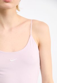 Απαλό ροζ αμάνικο μπλουζάκι με λεπτές τιράντες και στρογγυλή λαιμόκοψη. Διαθέτει μικρό λευκό λογότυπο Nike στο μπροστινό μέρος, κατασκευασμένο από μαλακό, ελαστικό υλικό.