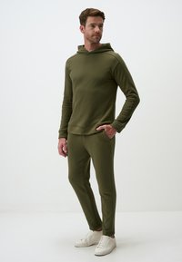 Olivenfarbener Hoodie und Hosen-Set aus weichem Stoff, ausgestattet mit einer Kapuze, gerippten Bündchen und seitlichen Taschen, kombiniert mit weißen Sneakers.