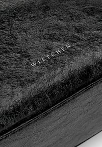 Černý kožený doplněk s texturovaným povrchem a jemným prasklým vzorem, s embosovaným logem "WITTCHEN" v stříbrné barvě.