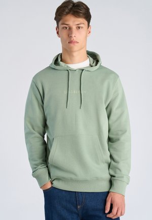 Young man with light brown hair wearing a light green hoodie with front pocket and dark blue jeans, standing against a plain light background.