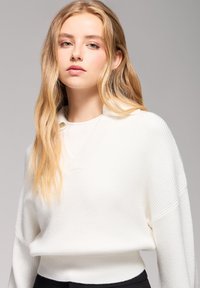 Biała sweter wykonany na drutach z dekoltem w serek i prążkowaną strukturą. Charakteryzuje się luźnym krojem i opadającymi ramionami. W połączeniu z bluzką z okrągłym dekoltem pod spodem.