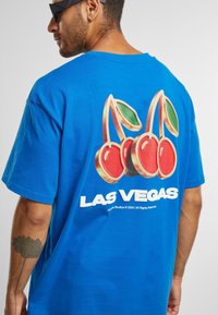 Mėlynas medvilninis marškinėlis su raudonų vyšnių ir žalių lapų grafika, ant nugaros dideliais baltais žodžiais užrašyta "LAS VEGAS".