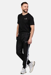 Zwart T-shirt met korte mouwen en zwarte trainingsbroek met zijstreep. De trainingsbroek heeft een elastische tailleband en logo-detail. Witte sportschoenen.