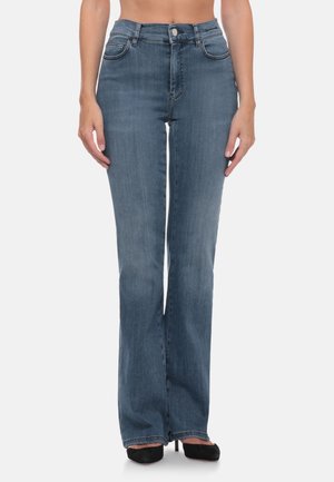 Donna che indossa jeans blu a vita alta a zampa e tacchi neri a punta, in piedi davanti a uno sfondo chiaro e uniforme.