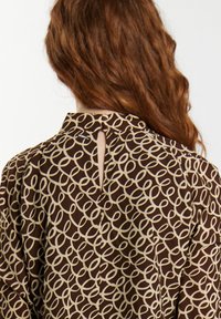 Eine rothaarige Person von hinten gesehen, die eine braune Bluse mit einem beigen Schlaufenmuster trägt und eine kleine Öffnung im Rücken nahe dem Hals hat.