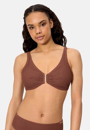 Mujer con cabello rizado que lleva un top de bikini marrón texturizado con un broche dorado en el frente, sobre un fondo liso de color gris claro.