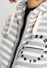 Persona che indossa una felpa con zip grigio e bianco a righe, con ricamo "Superdry Bronx New York 1991" e laccetti color crema.