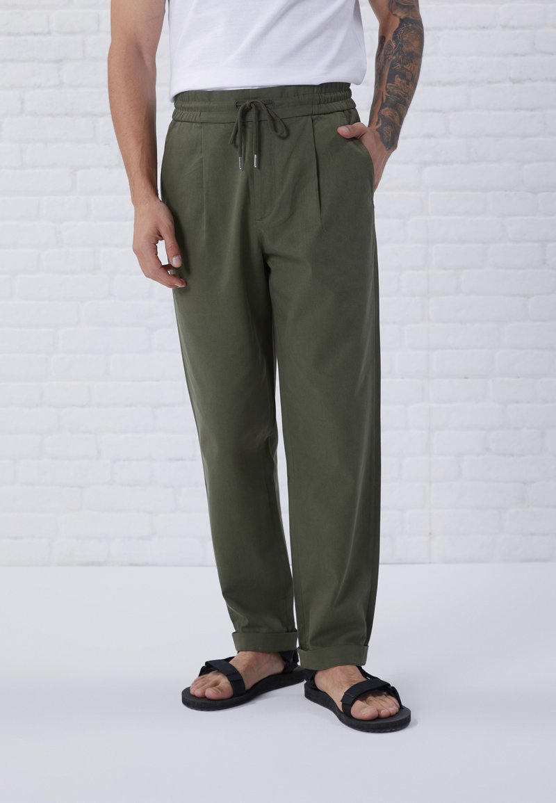 Pantaloni verde oliva con vestibilità rilassata, cintura elastica, tasche laterali e vita plissettata. Abbinati a sandali neri su sfondo bianco.