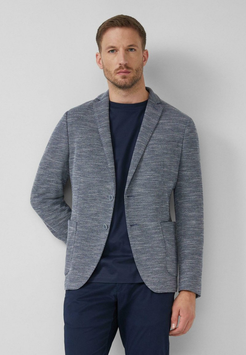 Grauer gewebter Blazer mit Revers, zwei Knöpfen und seitlichen Taschen, über einem marineblauen T-Shirt mit strukturiertem Finish getragen.