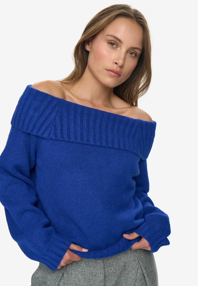 Pull bleu à épaules dénudées avec un col et des poignets côtelés, fabriqué dans un tissu doux et texturé ; associé à un pantalon gris à motifs.