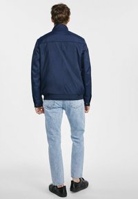 Granatowa kurtka z wysokim kołnierzem, wykonana z gładkiego materiału. Posiada ribbowe wykończenia na dole i przy mankietach, noszona z jasnoniebieskimi dżinsami i czarnymi butami.