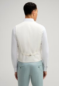 Weißer, strukturierter Weste mit einer Schnalle auf der Rückseite, getragen über einem hellblauen Anzug. Das Model zeigt mit dem Rücken zur Kamera und hebt das Design der Weste hervor.