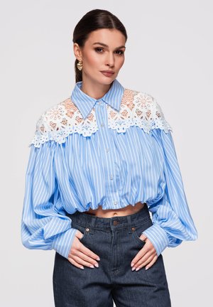 Kobieta w niebiesko-białej prążkowanej krótkiej bluzce z białą kwiatową koronką na karczku, bufiastymi rękawami, złotymi kolczykami i ciemnych jeansach z wysokim stanem.