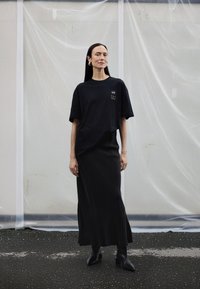 Žena s dlouhými tmavými vlasy, která nosí černé oversized tričko, černou dlouhou sukni a černé kozačky na podpatku, stojí na asfaltu před bílým průsvitným pozadím.