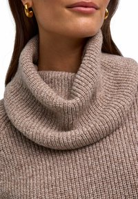 Maglione beige a collo alto con coste, dal tessuto morbido e colletto ampio e ripiegato. Orecchini a cerchio dorati visibili nell'immagine.