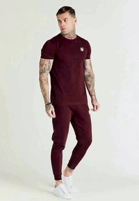 Conjunto atlético em bordeaux, incluindo uma t-shirt ajustada e calças de jogging ajustadas, feito de um tecido suave, com detalhes em branco no logotipo.