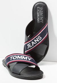 Sandales slides noires avec des brides bleu marine, rouge et blanche croisant le pied, avec les textes "TOMMY" et "JEANS" sur les brides et la semelle.