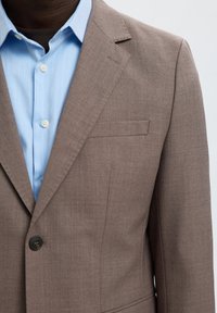 Gros plan d'une personne portant un blazer marron clair sur une chemise bleu clair avec des boutons blancs sur un fond neutre.