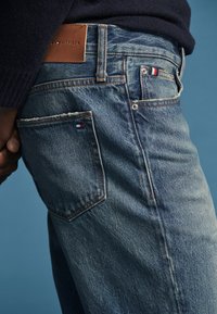 Κοντινό πλάνο ενός ατόμου που φορά μπλε τζιν Tommy Hilfiger με καφέ δερμάτινο λογότυπο και μικρές λεπτομέρειες λογότυπου στις τσέπες.