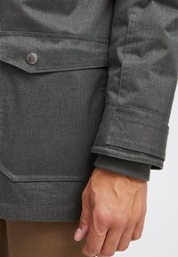 Pilkas herringbone tipo švarkas turi detales: kišenę su mygtuku, rankovę su raštiniu tekstūru ir lygią medžiagą su matomomis siūlėmis.