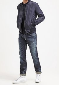 Marinblå bomberjacka med dragkedja, tillsammans med mörka denimjeans och vita högblänkta sneakers. Subtil textur och bleka mönster på jeansen.