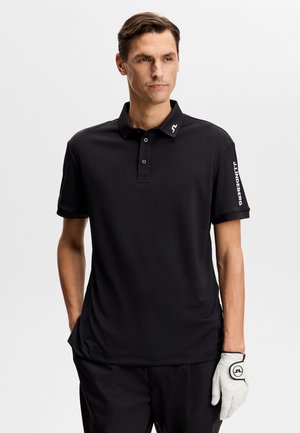 Man wearing black polo shirt with J.Lindeberg logo on sleeve and collar, black pants, and a white golf glove on left hand.