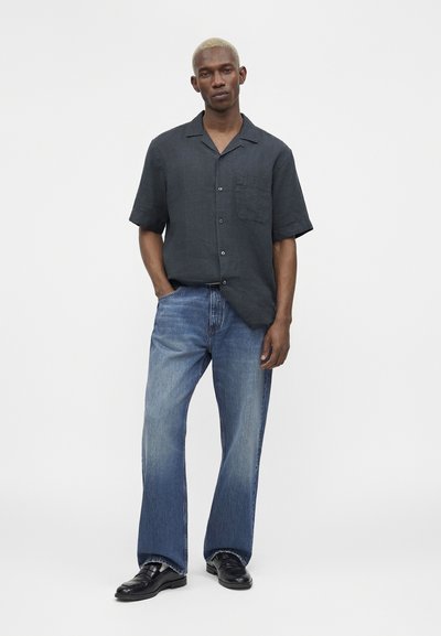 Άνδρας στέκεται με τα χέρια στις τσέπες, φοράει κοντομάνικο σκούρο πουκάμισο με κουμπιά, μπλε τζιν και μαύρα δερμάτινα παπούτσια σε λευκό φόντο.
