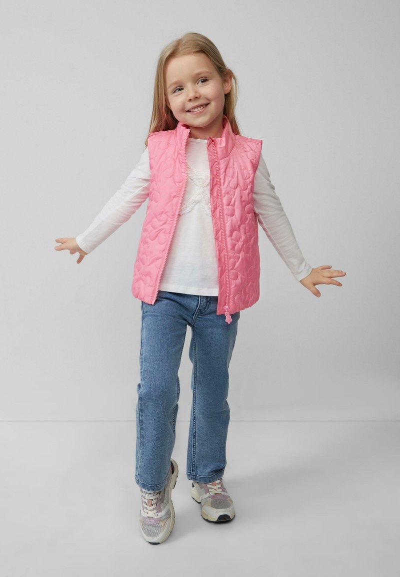 Rosa gesteppte Weste mit hohem Kragen, kombiniert mit einem weißen Langarmshirt und blauen Jeans. Besitzt einen Frontreißverschluss und sportliche Sneaker.