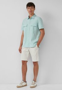 Hellgrünes kurzärmliges Hemd mit zwei Brusttaschen, Button-down-Kragen, getragen mit cremefarbenen Shorts und beigen Sneakern. Sanfte Stoffstruktur.