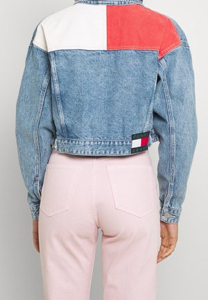 Person iført en cropped denimjakke med røde og hvide farveblokke på øvre del af ryggen og lyserøde bukser, stående mod en ensfarvet baggrund.