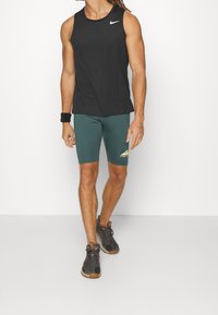 Homme portant un débardeur noir Nike, un short Nike vert foncé, un bracelet noir et des chaussures de sport, se tenant devant un fond blanc.