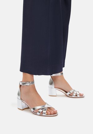 Ezüst sarkú szandál többutas keresztpántokkal, bokapántos záródással és tömbsarkú dizájnnal, navy kék nadrággal párosítva.
